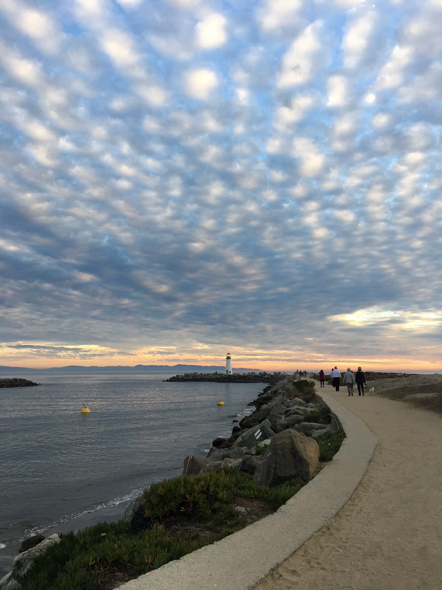 A gravel path leads along the bay to a lighthouse in the distance; the sky above is full of clouds as the sun sets.