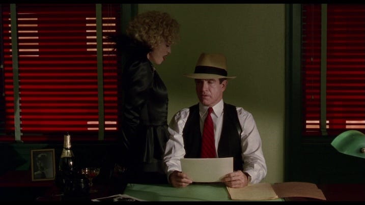 Warren Beatty as Dick Tracy sits at his desk reading a paper while Madonna’s character, Breathless Mahoney, stands beside him—bathed in green and red lighting.