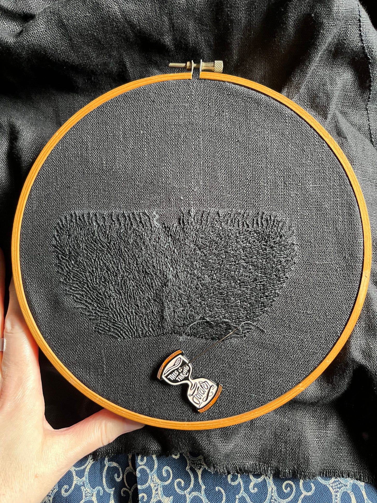 Lia’s fingers show a wooden embroidery hoop with a 4.5 by 5 inch trapezoid of black on black whipped backstitch that looks a bit like flames, but if you’ve been following this piece you’ll know it’s pubic hair. It’s definitely pubic hair. There’s a white chalk line on the upper edge showing about a centimetre in height to go, especially in the centre couple inches, and some chalk lines within the hairs. An hourglass-shaped needle minder that reads “this is taking forever” is attached to the cloth.  