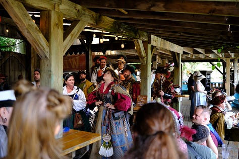Bards singing in a tavern wearing medieval garb, an elf and wizard surveying goods at a shop, fire-twirler show, Geralt and another warrior deep in conversation, a man playing dead with an arrow under his armpit, guests walking toward the camera, a woman with a horse head looking over a table.