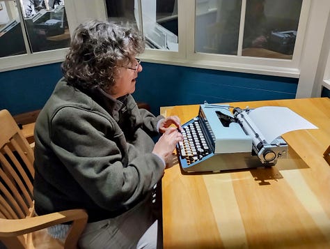 Desk with wooden chair and typewriter in mock office; Annette typing;corner of typewriter with Annette's typed note
