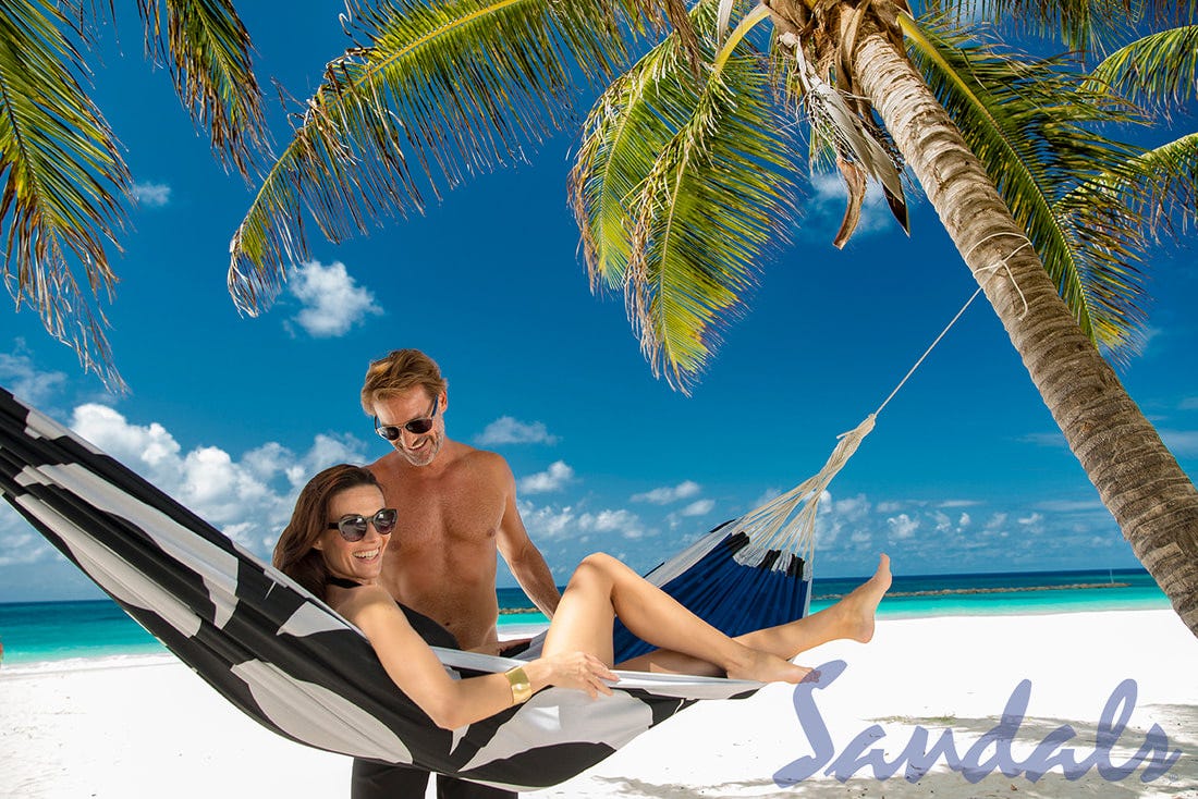 Promotional photo for Sandals Resorts depicts a woman in beachside hammock with a smiling, shirtless man standing and smiling beside her.