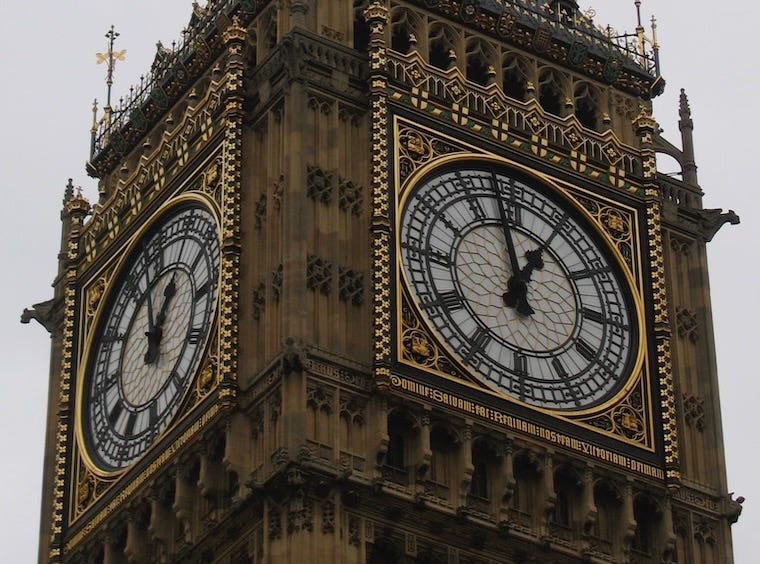 Close-up view of two clock faces on the Elizabeth Tower, commonly known as Big Ben, in London. The ornate clock faces are framed in gold and black detailing, with the time showing 12:58. The Gothic architectural details of the tower are visible, along with a cloudy grey sky in the background.