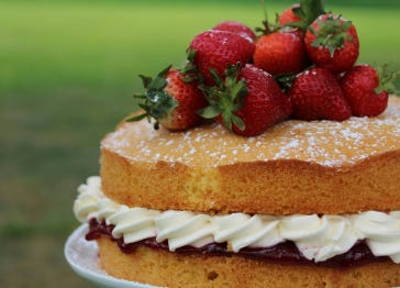 Photo shows a sponge cake with red jam and white cream filling, topped with strawberries, in an outdoor setting with green behind, by balesstudio on Unsplash
