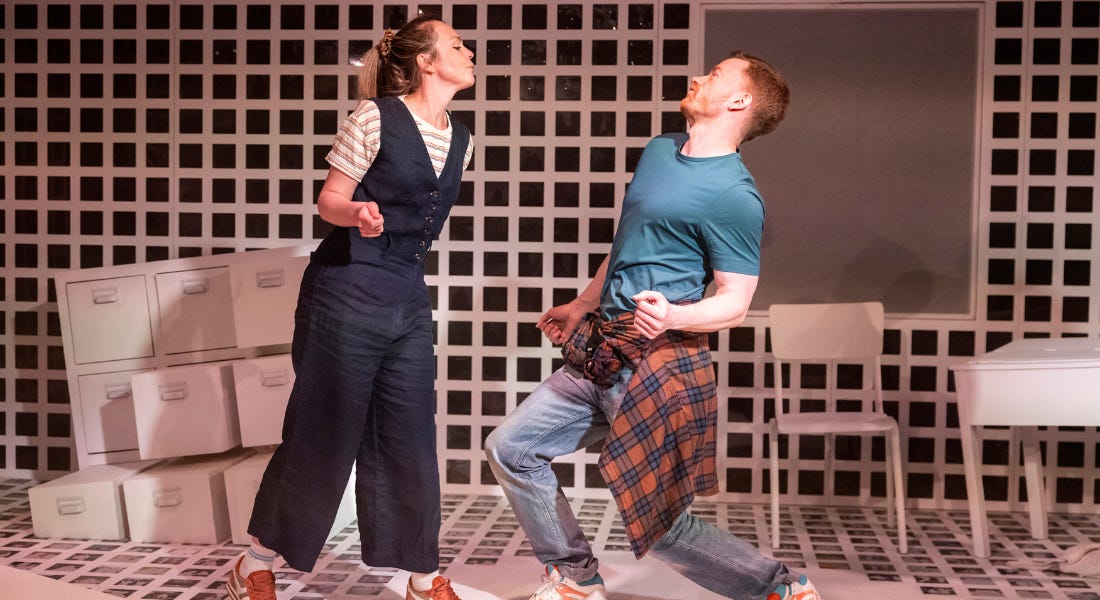 A woman wearing a blue jumpsuit and stripy t-shirt and a man wearing a green t-shirt and jeans are dancing together. She leans towards him and he leans backward. In the background is a room with a white grid pattern on the walls and floor and white furniture which slants into the floor. A woman wearing a blue jumpsuit and stripy t-shirt and a man wearing a green t-shirt and jeans are dancing together. She leans towards him and he leans backward. In the background is a room with a white grid pattern on the walls and floor and white furniture which slants into the floor.