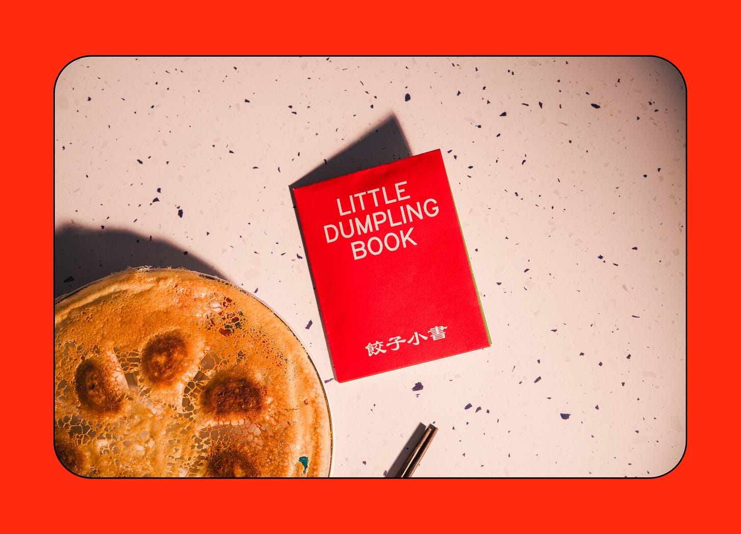A red book titled "Little Dumpling Book" lies on a speckled white surface next to a partially visible pan-fried dumpling dish and a pen.