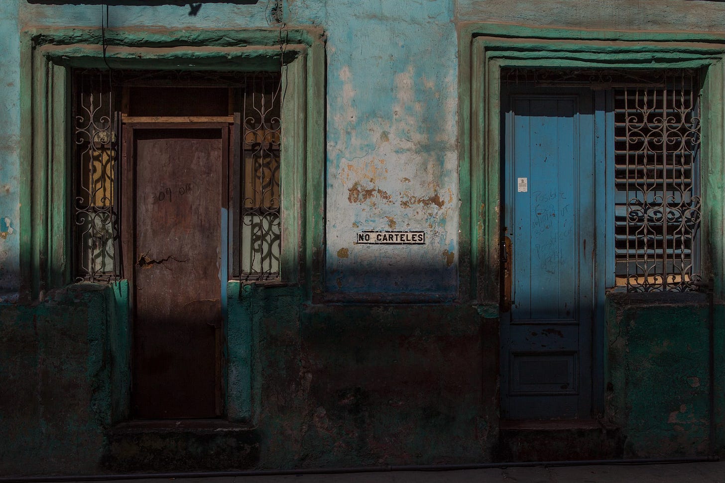 A weathered turquoise wall in Havana features two old doors — one boarded, one bright blue — framed by ornate metal bars. In the center, a small sign reads “NO CARTELES” (No posters). Soft, late afternoon light cuts across the scene, revealing decades of wear and subtle graffiti.