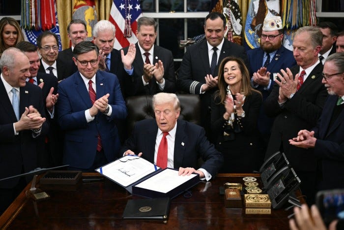 Donald Trump sits at a desk in the Oval Office displaying a signed bill as a group of people around him applaud