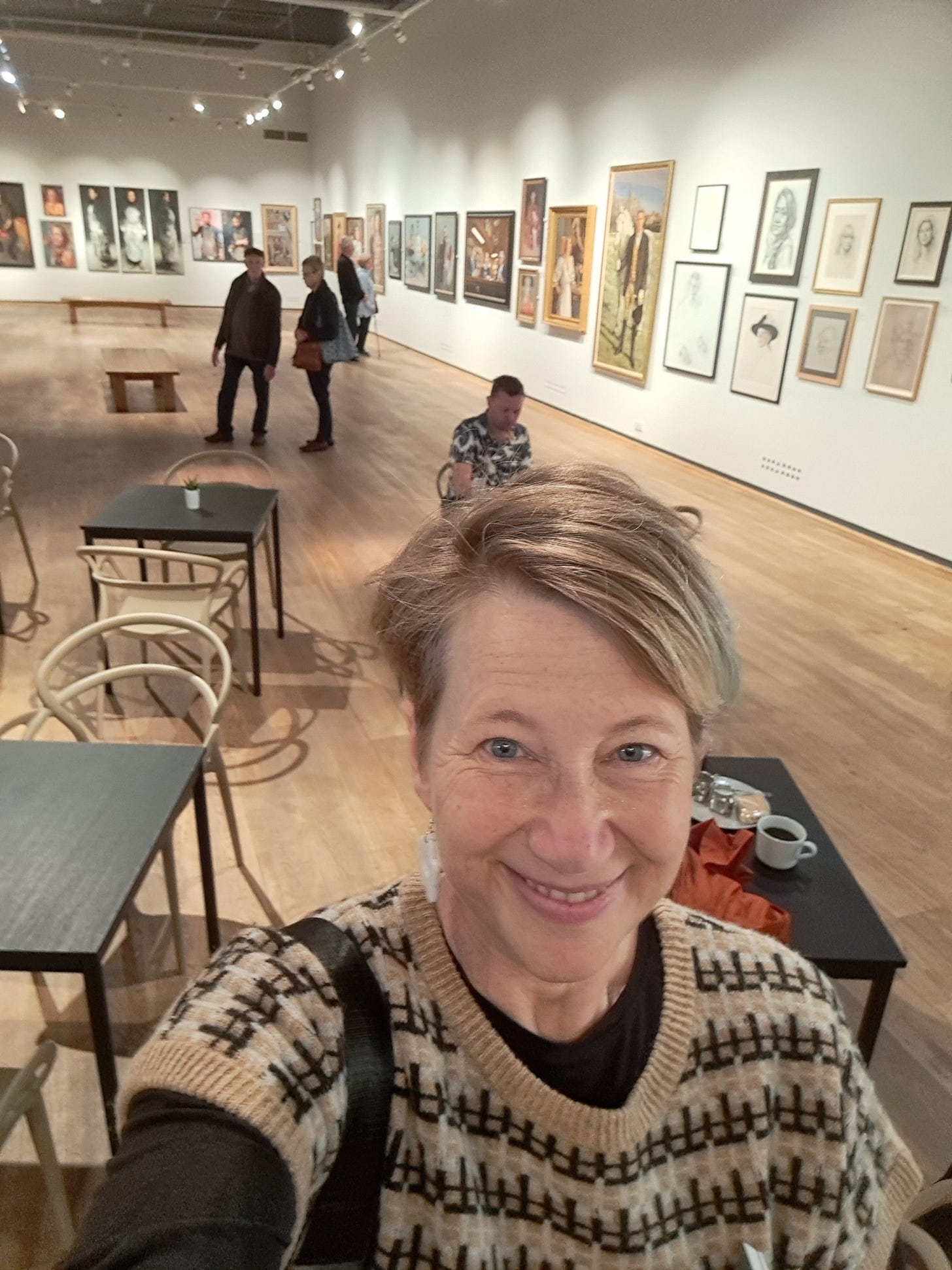 Gail looking up at the camera, in the main hall of the Mall Galleries. Pictures lining the walls, cafe tables and people