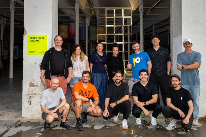 A group of people standing and kneeling in front of a building with large open doors. Visible individuals include Wenke Blüt, Amy Goodchild, Alexis André, Monica Rikic, Niklas Roy, Yose Llobet, Glitch Life, Heeey Eth, Rotor Studio, Xavier Hébert-Baggott, Bustavo, and Julia Hespeneide. A yellow sign with black text reading "REFLECTIVE DREAMS" is on the left wall.