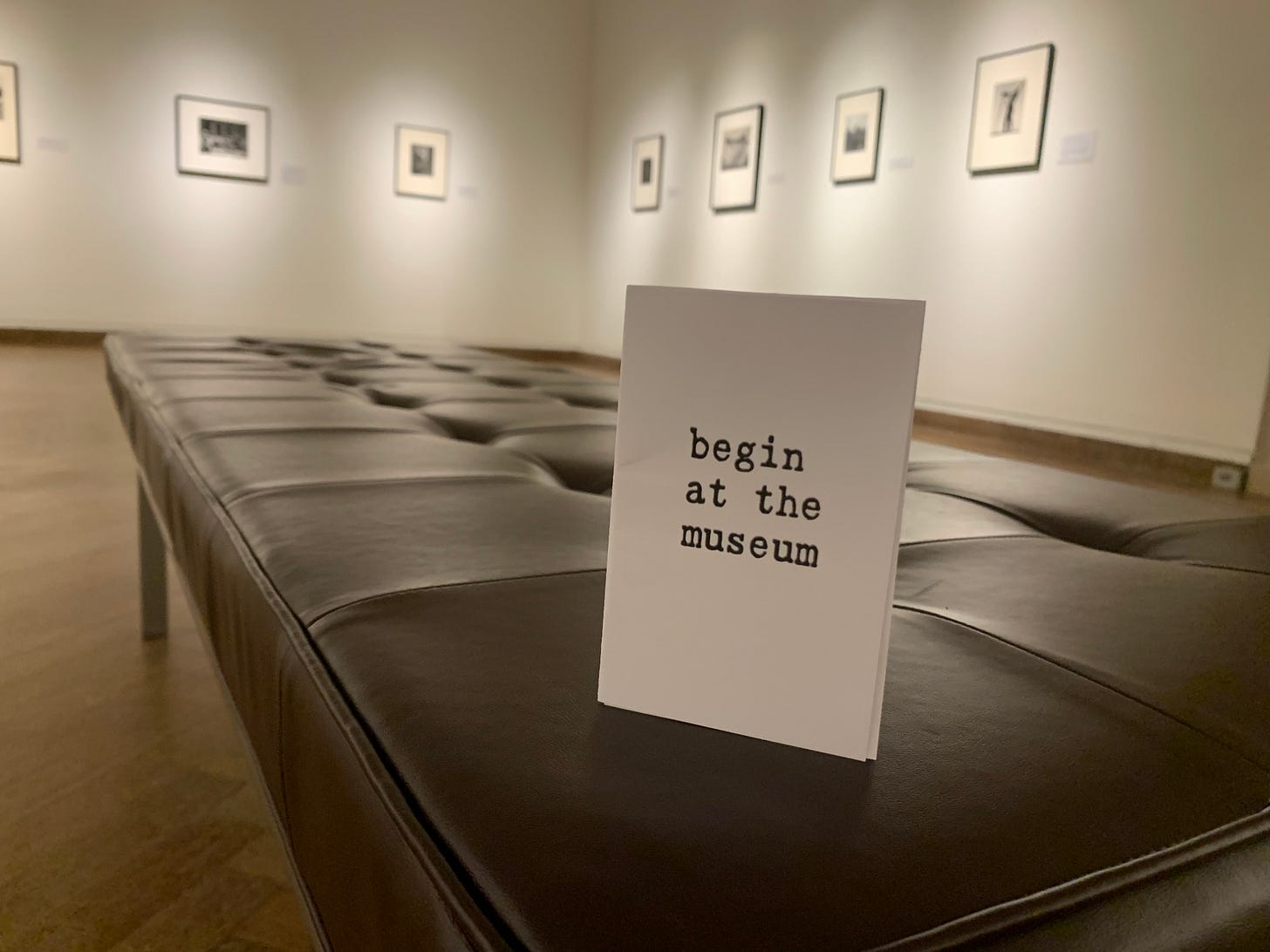 A zine titled "Begin at the Museum" rests on a bench in an art museum, with a row of framed images visible behind it