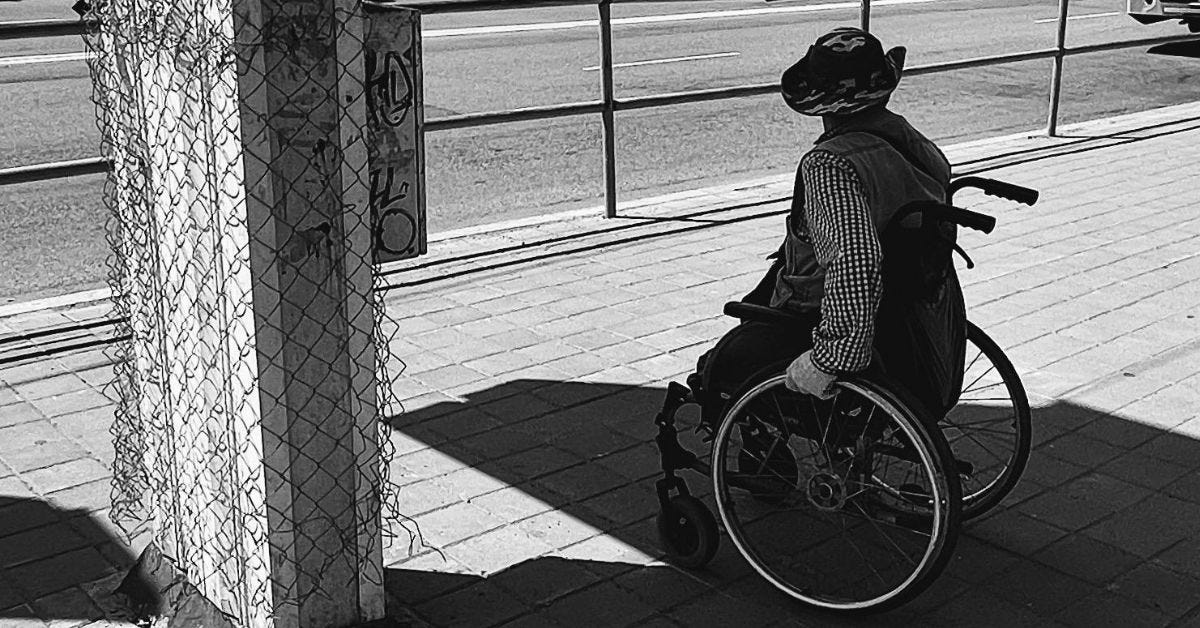 Black-and-white photo of a person in a bucket hat and vest, seen from behind, wheeling a manual wheelchair along a brick sidewalk beside a road, past a concrete pillar wrapped in torn chain-link fencing.