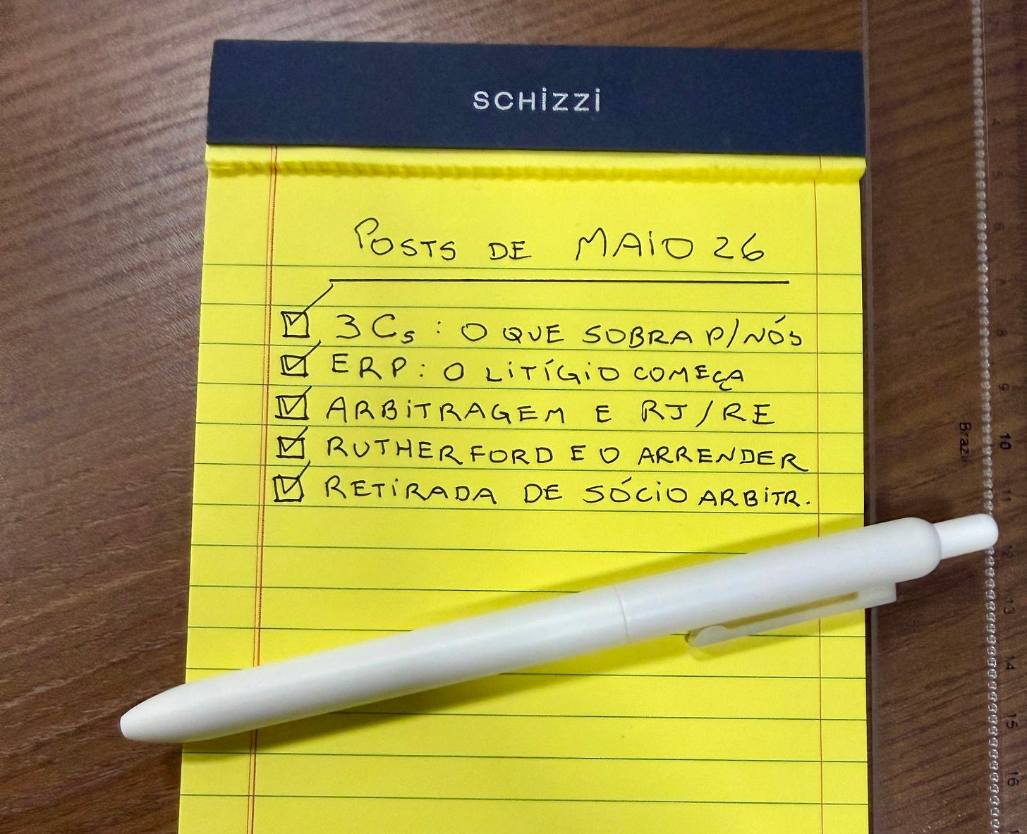 Bloco de anotações amarelo da marca Schizzi sobre mesa de madeira, com caneta branca apoiada. Em caligrafia manuscrita, o título "Posts de maio 26" e cinco itens marcados com check: "3 Cs: o que sobra p/ nós"; "ERP: o litígio começa"; "Arbitragem e RJ/RE"; "Rutherford e o aprender"; "Retirada de sócio arbitr.".