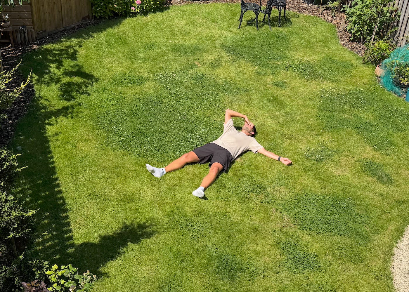 The author lies spread out on a sunny garden lawn, with one hand covering his face, surrounded by grass and garden plants.