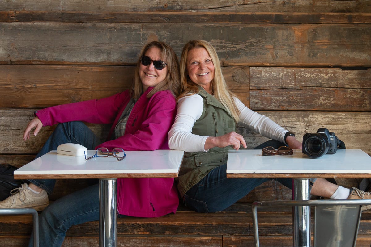 Two smiling women sitting back-to-back at a café table, one with a camera and the other with glasses and a case on the table.