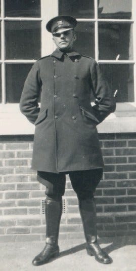 A black and white photo of a police officer in motorcycle gear. He wears knee length leather boots over puffy trousers and a long thigh length coat with a hat.