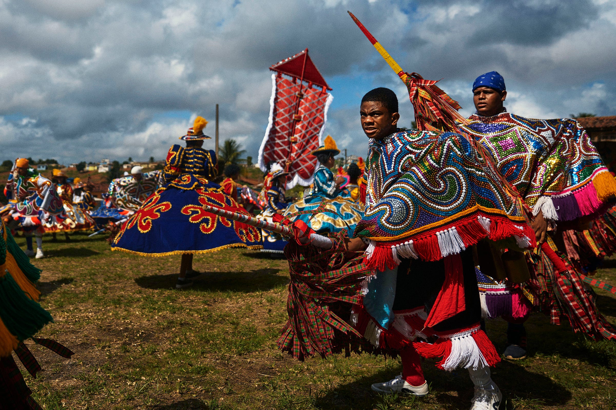 People in colorful, ornate costumes perform a traditional outdoor dance on a grassy field under a cloudy sky. Two men in the foreground hold decorated sticks, while others dance in the background with flowing capes and bright headpieces.