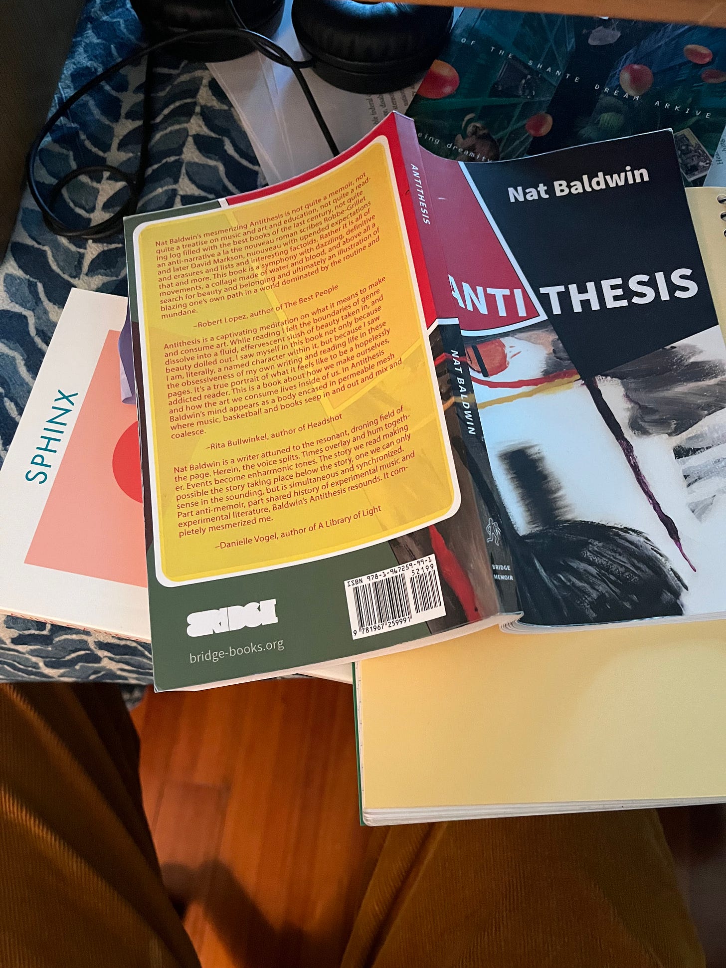 An image of nat baldwin's antithesis splayed open on top of the books Sphinx and Shante Dream Arkive, also part of the tableaux are printed pages, headphones, and a blue and white patterned tablecloth.