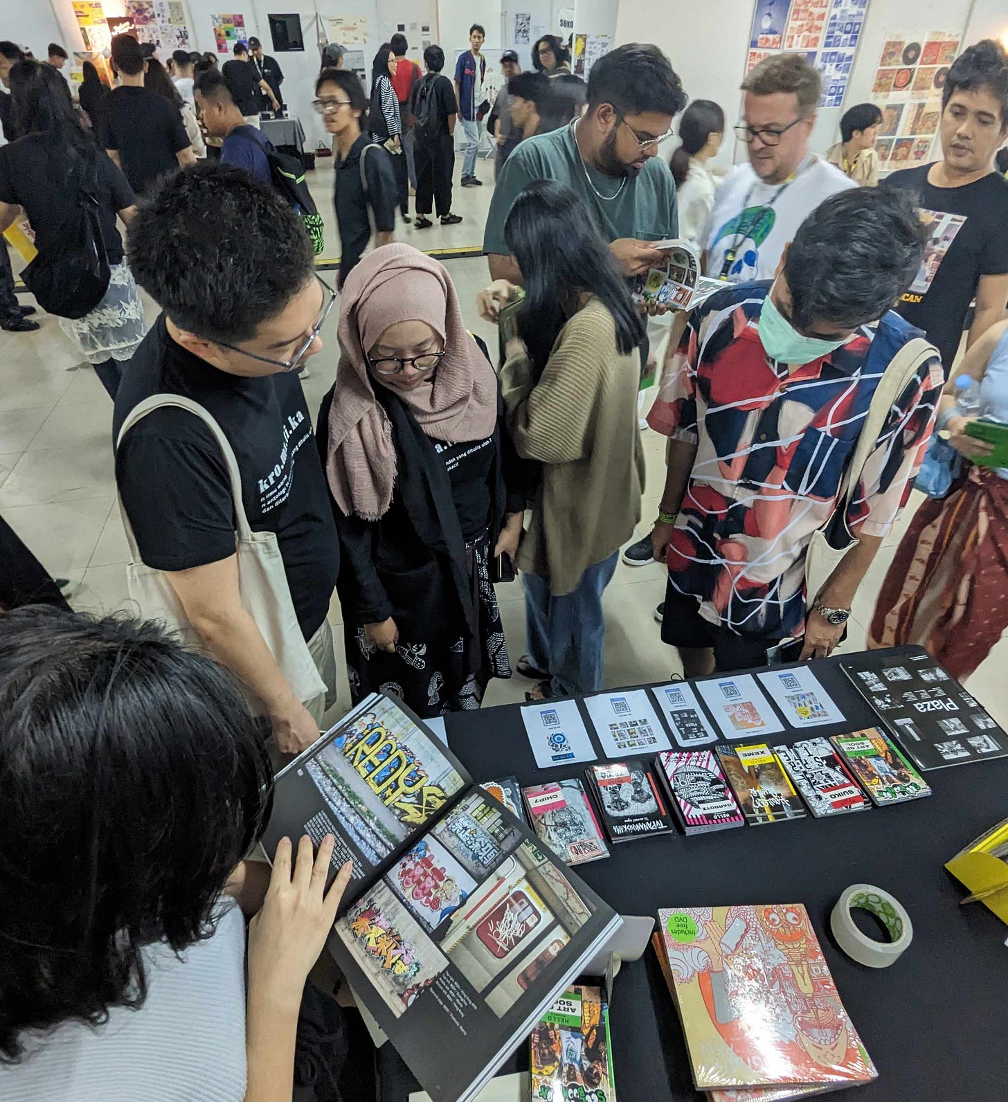 In this image, a group of people at the Jakarta art book fair in 2023 are gathered around a booth displaying various books and zines from Soi Books and Stickerbomb World. One person is sitting and flipping through a graffiti art book, showing colorful and vibrant street art pieces. On the table, there are several books, zines, and sticker packs neatly arranged, with QR codes for more information. The attendees, including a woman wearing a hijab, are engaged in exploring the booth, while the busy fair in the background shows other participants and booths in an active and lively environment.