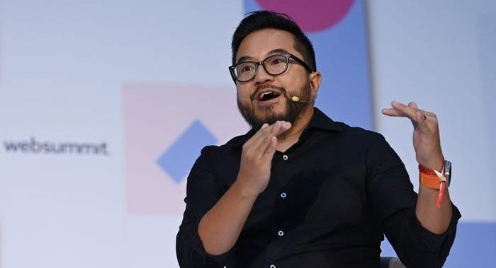 Garry Tan, the CEO of Y Combinator, is gesticulating with his hands and is mid-speech. He is wearing a smart watch with an orange band, a black collared button up, and blue jeans. He is sitting in a grey chair in front of a background with text that reads, "websummit."