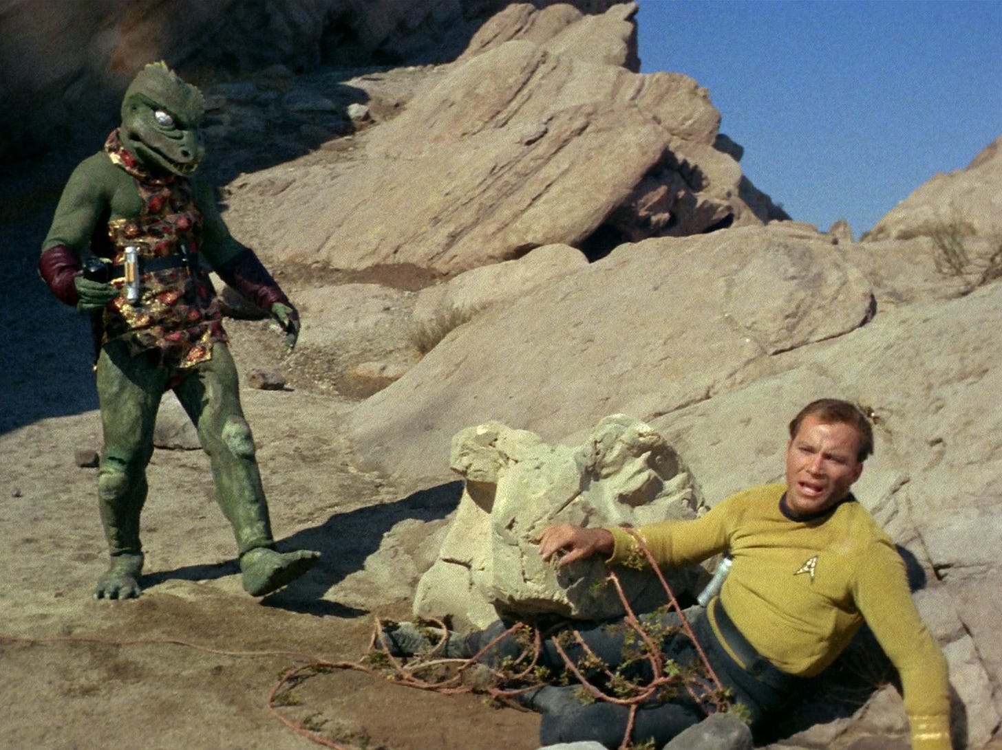 Captain Kirk is on the ground, with vines tangled around his legs, as a green reptilian alien approaches him menacingly