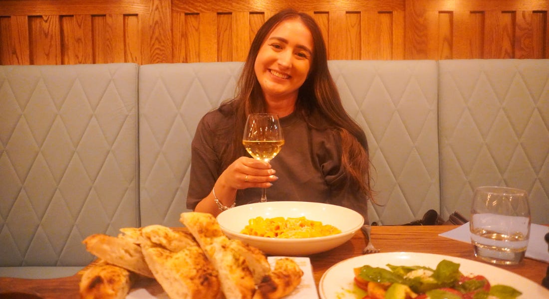A woman - Eve Smallman - sitting at a table holding a glass of wine with bread, salad, and pasta in front of her A woman - Eve Smallman - sitting at a table holding a glass of wine with bread, salad, and pasta in front of her