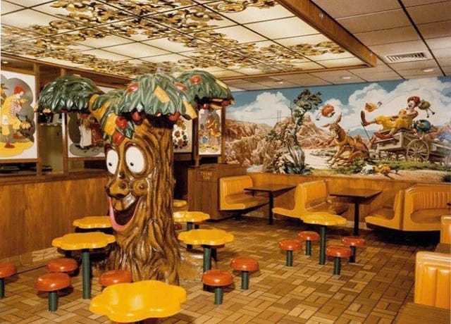 A 1970s-era McDonald’s interior featuring a whimsical, anthropomorphic fiberglass tree with wide cartoon eyes and leafy canopy. Surrounding it are orange-yellow mushroom‑style stools and round tables amid brown-toned tile flooring and booths, evoking a kitschy, playful fast‑food dining space. A 1970s-era McDonald’s interior featuring a whimsical, anthropomorphic fiberglass tree with wide cartoon eyes and leafy canopy. Surrounding it are orange-yellow mushroom‑style stools and round tables amid brown-toned tile flooring and booths, evoking a kitschy, playful fast‑food dining space.
