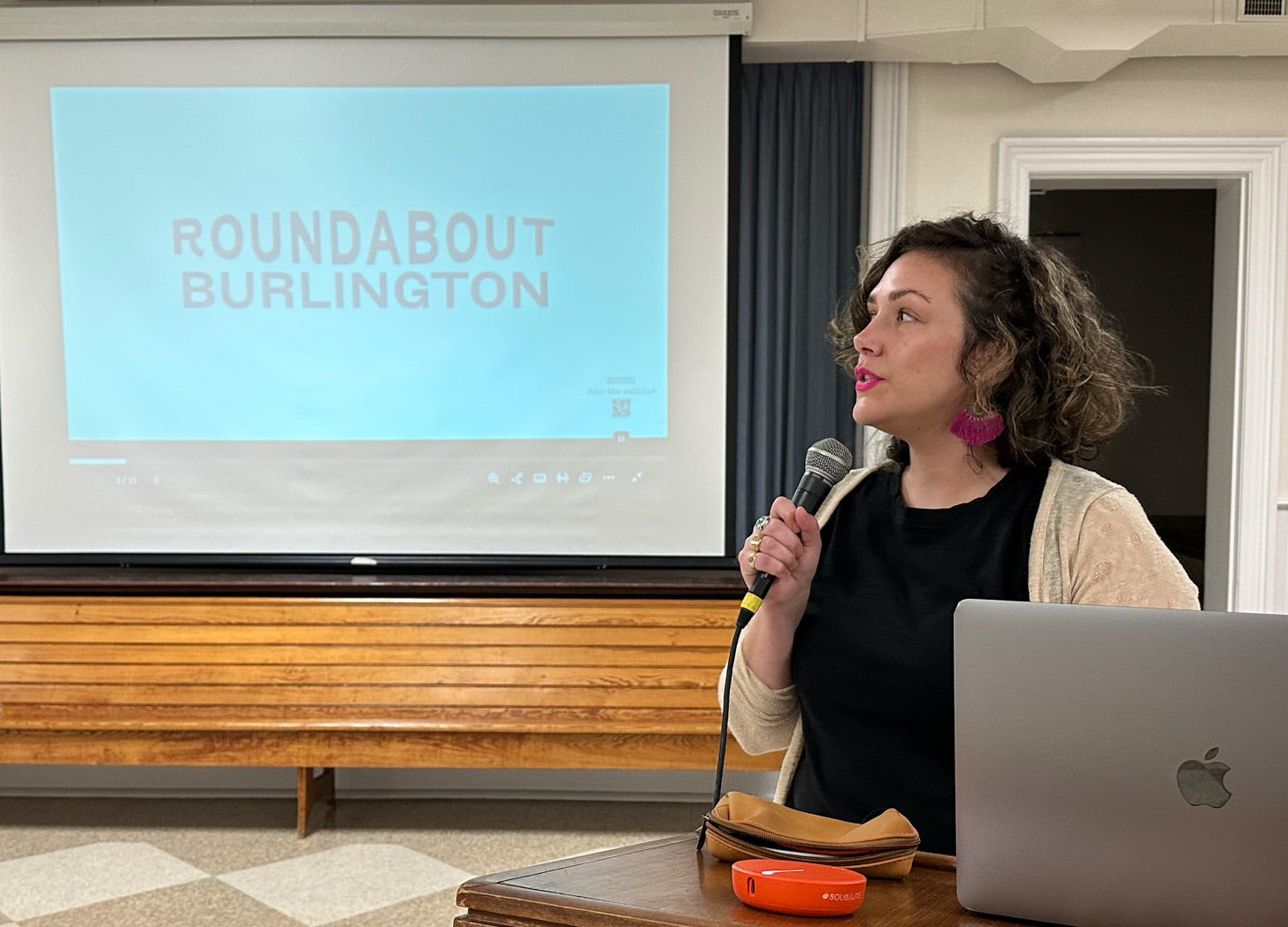 Presentation scene with a projector screen displaying "ROUNDABOUT BURLINGTON" in light text on a blue background. A woman speaks into a microphone, standing next to a MacBook laptop and an orange device, with wooden paneling and curtains in the background. Presentation scene with a projector screen displaying "ROUNDABOUT BURLINGTON" in light text on a blue background. A woman speaks into a microphone, standing next to a MacBook laptop and an orange device, with wooden paneling and curtains in the background.