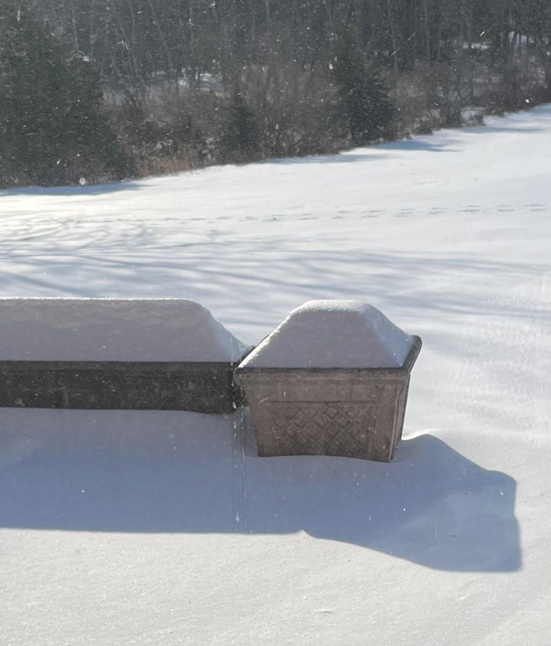 Photo of a snowy backyard including a snow covered patio wall and a snow covered planter taken through a dirty window.