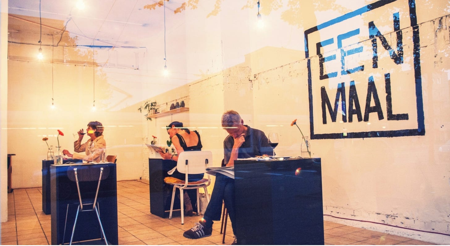 In a glassed-in shop, three diners sit at small black cube tables set for one person each. The words "Een Maal" are painted on a white wall of the space. In a glassed-in shop, three diners sit at small black cube tables set for one person each. The words "Een Maal" are painted on a white wall of the space.