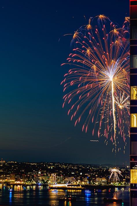 Night fireworks exploding beside Seaport skyscraper with Boston skyline and harbor reflections
