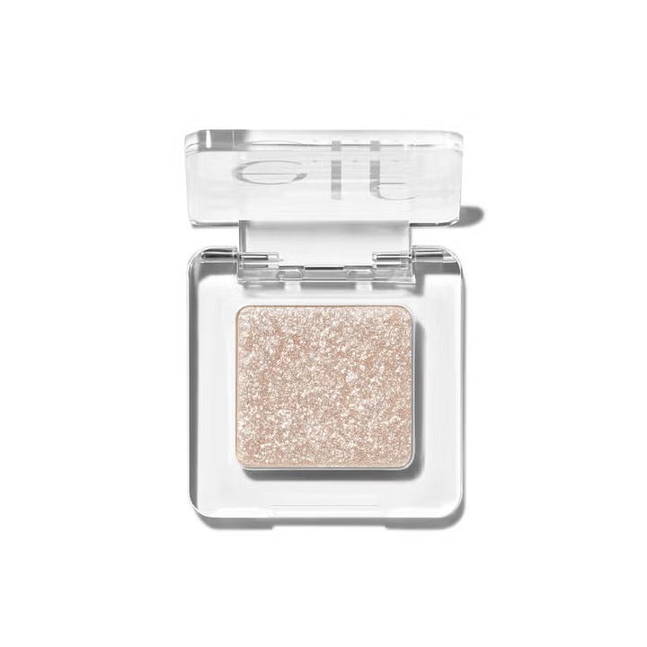 A close-up, top-down view of a square, clear acrylic makeup compact. Inside is a shimmering, pressed glitter eyeshadow in a pale champagne-gold shade with a high-shine, reflective finish.