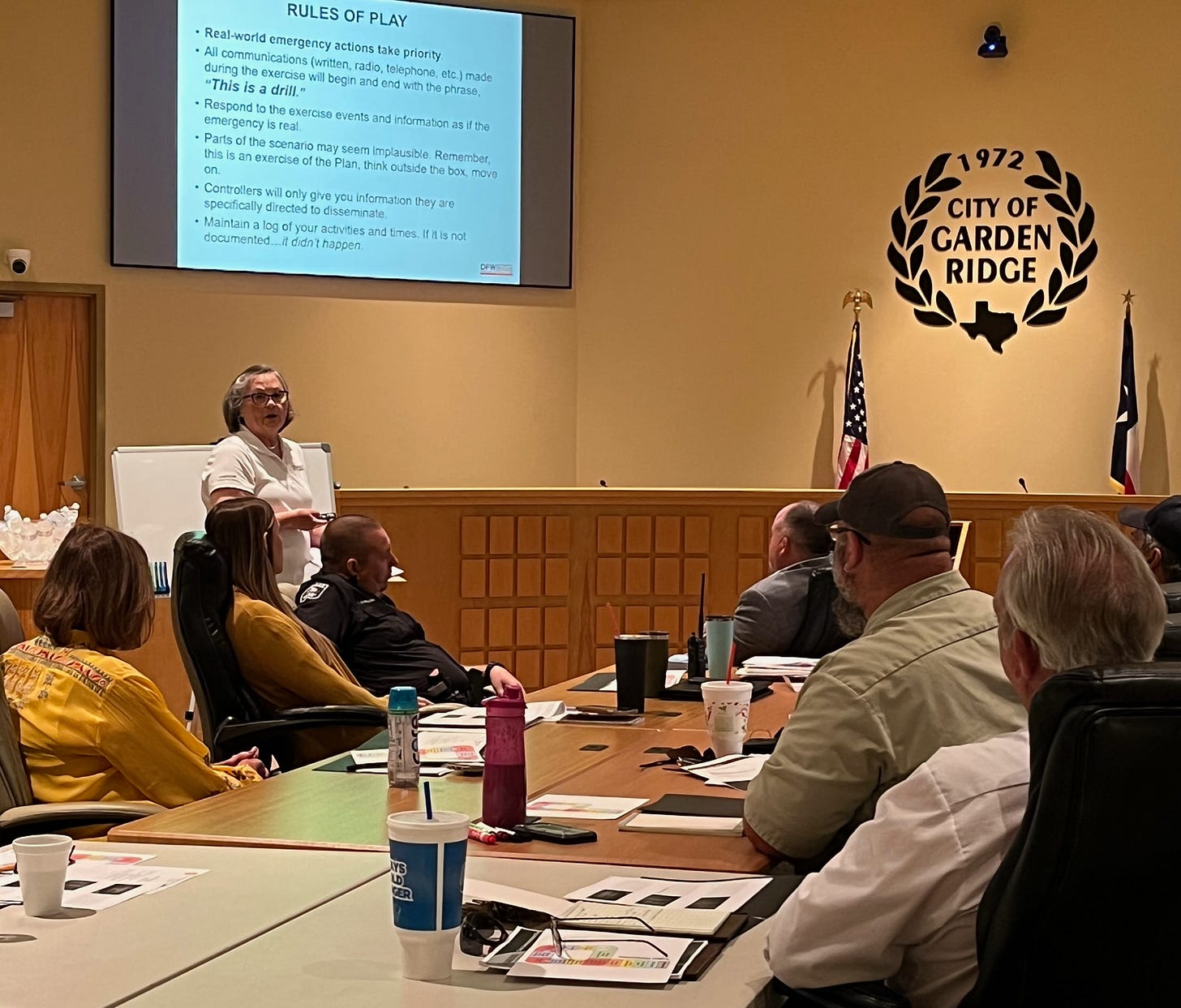 A woman stands at the front of a conference room in the City of Garden Ridge, Texas, leading a tabletop emergency exercise. Behind her, a large screen displays “Rules of Play” for the drill, including a bolded reminder: “This is a drill.” Attendees—including public safety personnel and city staff—are seated around a U-shaped table with printed materials, drinks, and notepads in front of them. The city’s seal and American and Texas flags are visible on the wall behind the speaker.
