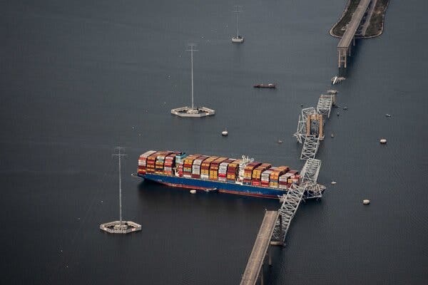 An aerial view of a large cargo ship, with the front of the ship sitting under a collapsed bridge. An aerial view of a large cargo ship, with the front of the ship sitting under a collapsed bridge.