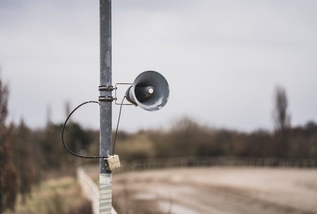 Photo of a loudspeaker attached to a metal pole on what appears to be a rural road. Photographer is Mika Baumeister and the photo is from Unsplash.