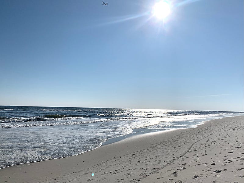 a photo of a beach, the waves are crashing on the sand, the sky is blue and cloudless with the sun bright and a small plane in the distance.