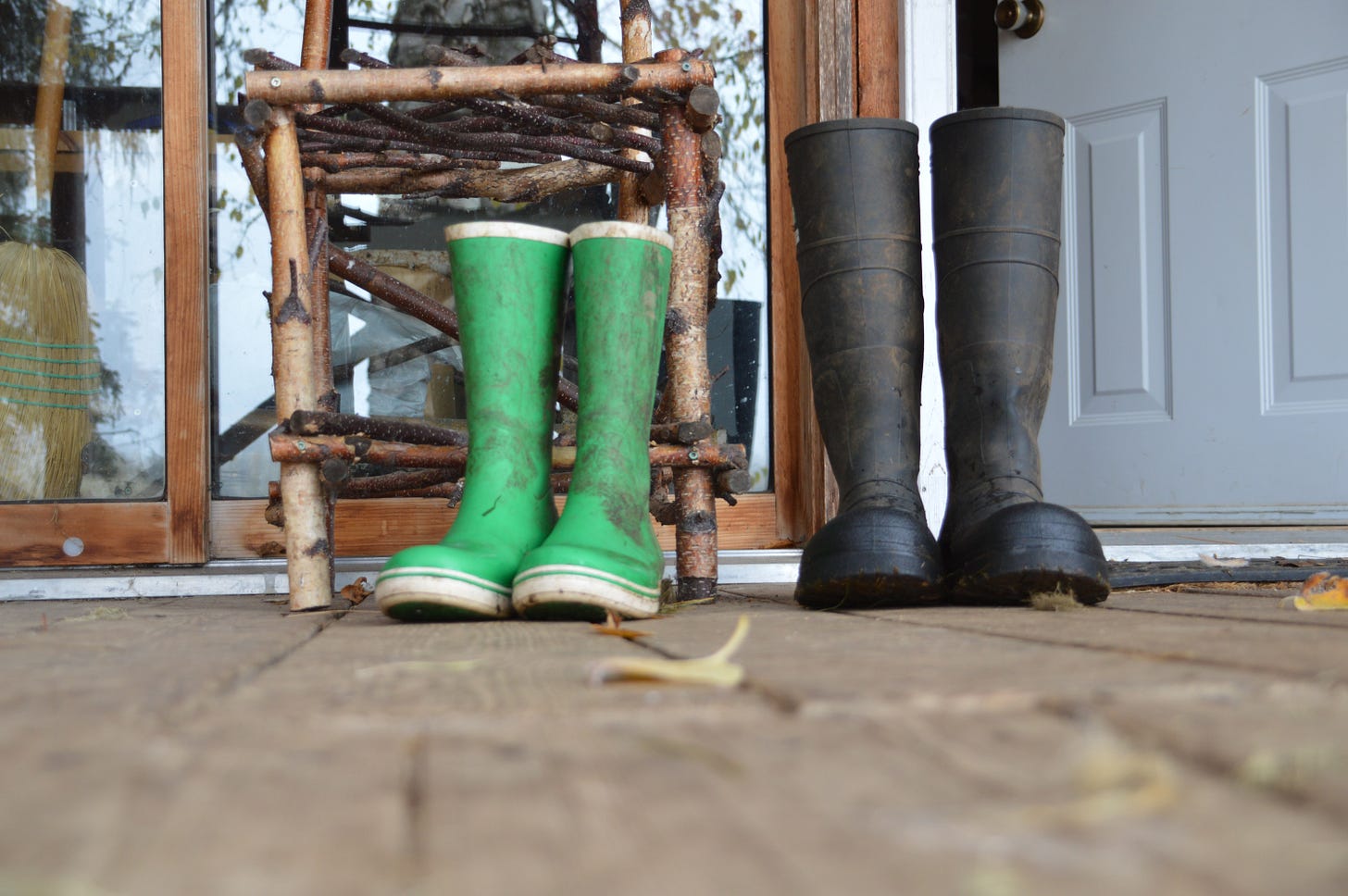 Two pairs of rubber boots - green and black - stand in front of a wooden chair, outside a doorway entrance Two pairs of rubber boots - green and black - stand in front of a wooden chair, outside a doorway entrance