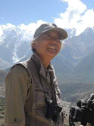A photo of a middle-aged Japanese man with binoculars around his neck and mountains in the distance. A photo of a middle-aged Japanese man with binoculars around his neck and mountains in the distance.