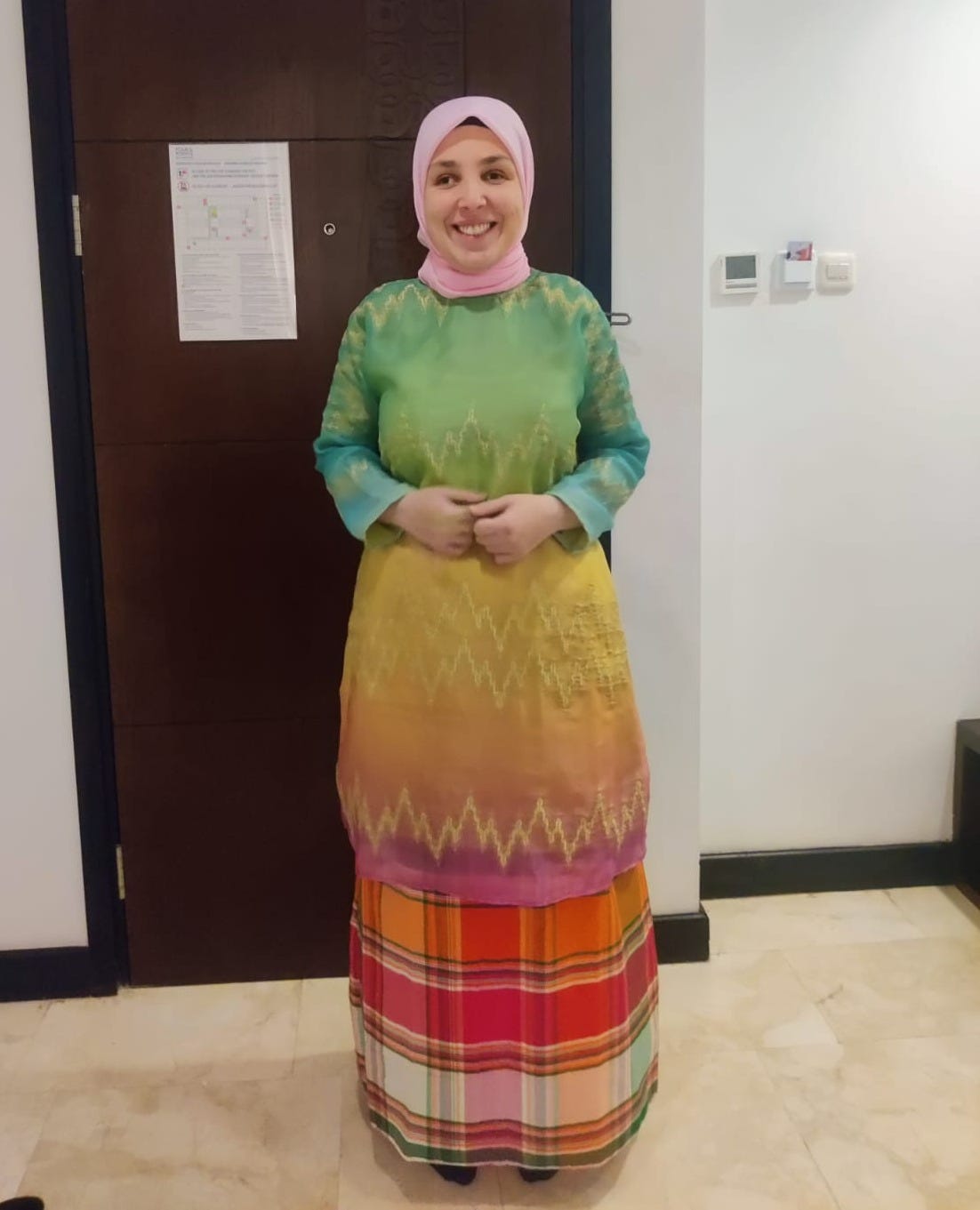 Sara stands, smiling wearing a brightly colored long sleeve garment. It is ombre, green at the top and fades into yellow, orange, and eventually pink on the bottom. She wears a longer, pink and orange plaid garment underneath, and a pink hijab. Sara stands, smiling wearing a brightly colored long sleeve garment. It is ombre, green at the top and fades into yellow, orange, and eventually pink on the bottom. She wears a longer, pink and orange plaid garment underneath, and a pink hijab.