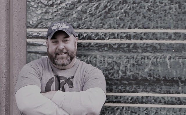Shannon, a white man with a beard, smiles at the camera. His arms are crossed and he's wearing a baseball cap. 