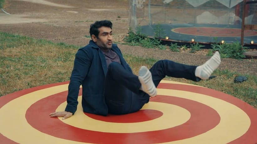 Kumail Nanjiani sits on a target with his legs lifted like he's giving birth