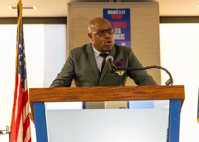 A bald Black man stands at a podium next to a U.S. flag wearing a grey suit and brown tortoise glasses A bald Black man stands at a podium next to a U.S. flag wearing a grey suit and brown tortoise glasses