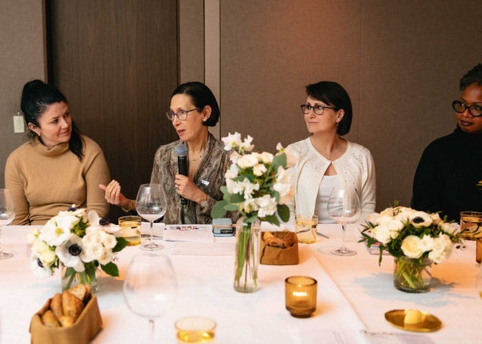 Three women at a long dinner formal dinner table listen to a fourth woman who is holding a microphone and speaking. Three women at a long dinner formal dinner table listen to a fourth woman who is holding a microphone and speaking.