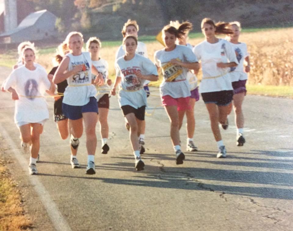 1990s photo of high school runners running on a rural road, tied together with loops of yellow rope. Among them is Laurie McGinley, second from the left in blue shorts giving it her stinking all.