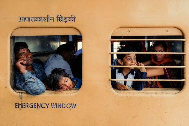 Depuis les fenêtres d'un train indien, on aperçoit un père visiblement épuisé, un enfant endormi, et deux autres enfants au regard joyeux, les yeux baissés vers le quai