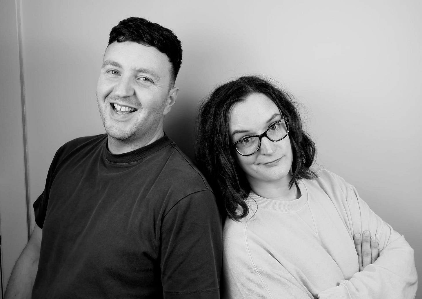 Ben and Mel in black and white. Mel looks quizzical and has her arms crossed.