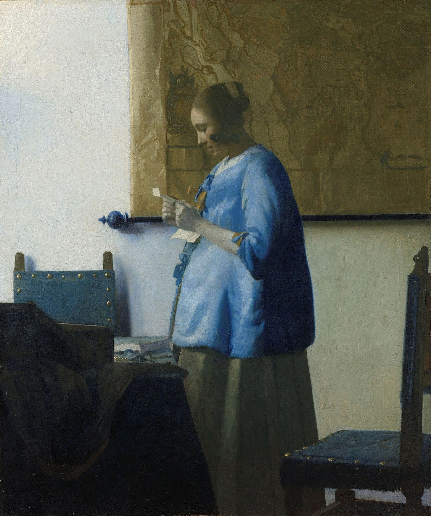A woman in a blue jacket stands in profile before a wall map and reads a letter held in both hands, with a second sheet on the table beside her A woman in a blue jacket stands in profile before a wall map and reads a letter held in both hands, with a second sheet on the table beside her