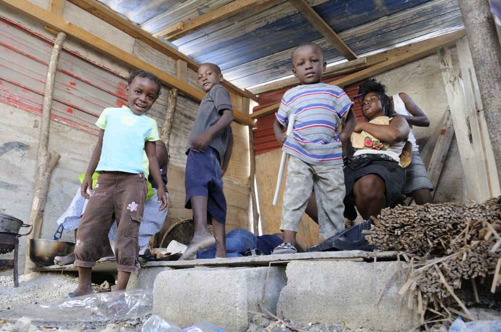 Children and adults stand and sit inside a fragile shelter made of wood and corrugated metal, with cooking pots and bundles of sticks on the ground. Three children face the camera in the foreground while two women, one holding a baby, sit behind them. The scene highlights the vulnerable living conditions of families affected by statelessness in the Dominican Republic.
