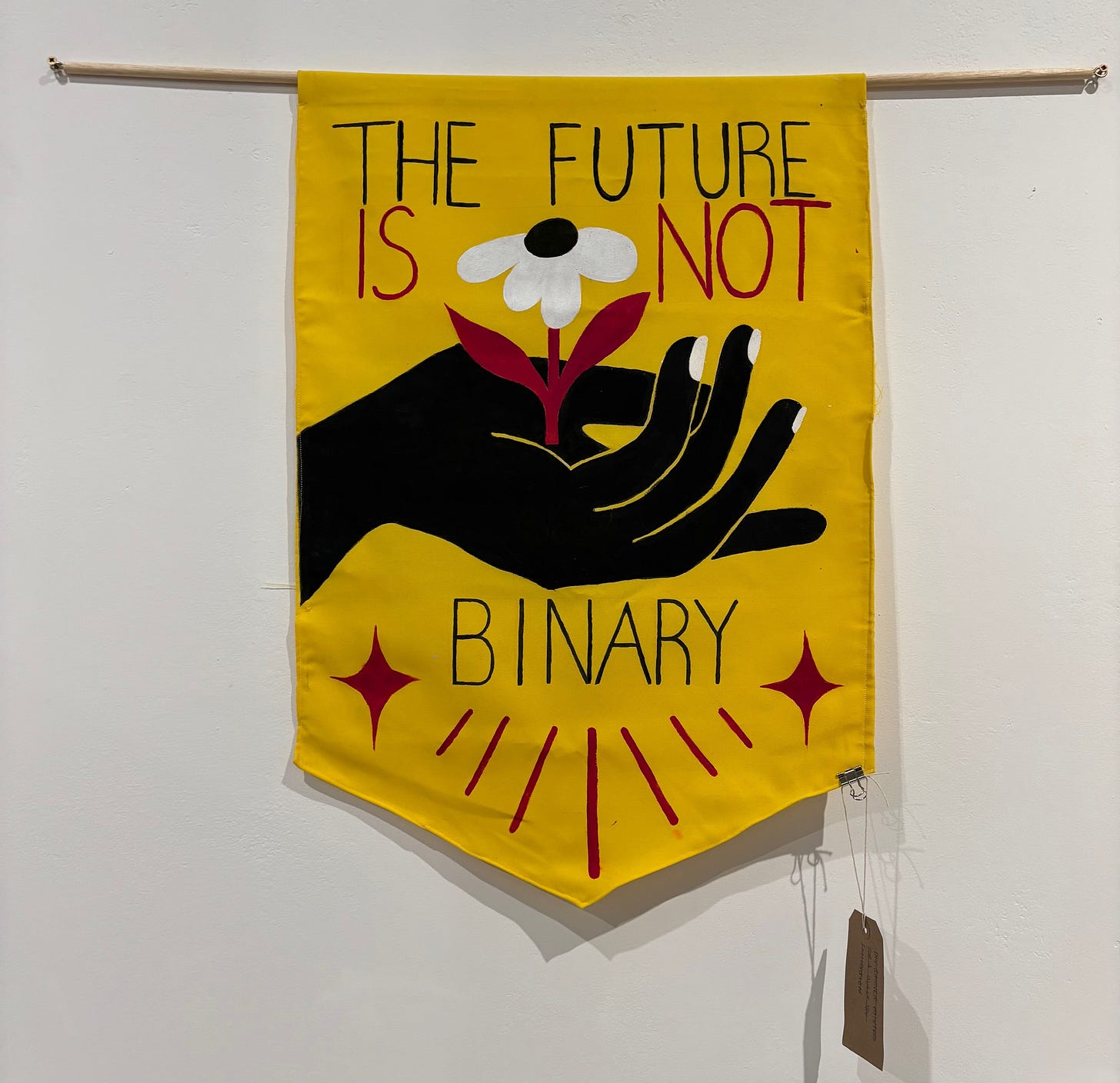 Yellow textile protest banner with black and red lettering that reads THE FUTURE IS NOT BINARY. In the centre a black hand cradles a white flower with red stem in an upturned palm