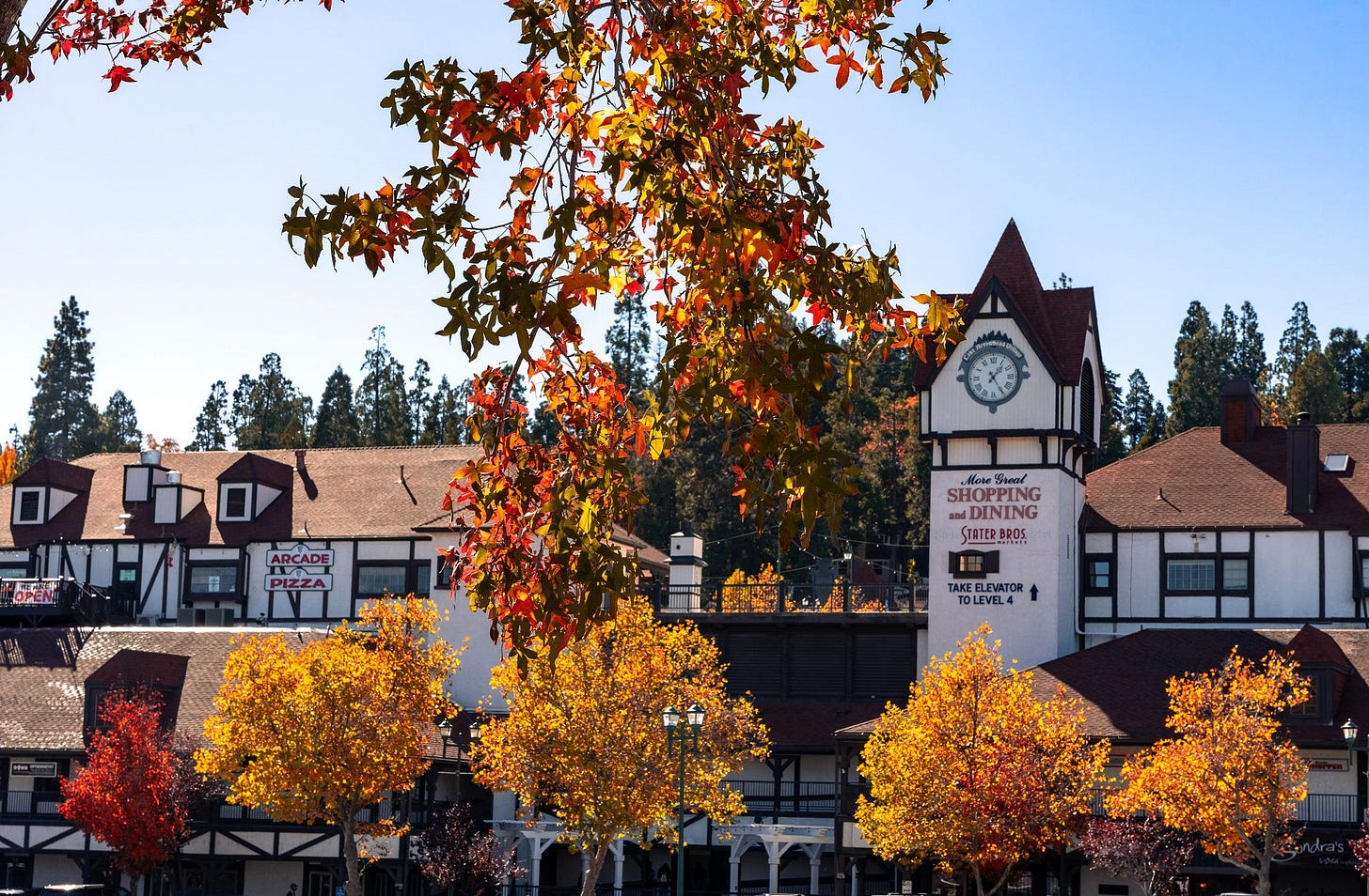 Lake Arrowhead Village in autumn, featuring Tudor-style buildings with dark timber framing and cream exteriors. A tall clock tower rises above storefronts advertising shopping and dining, while vibrant red, orange, and yellow leaves frame the scene against a backdrop of pine-covered hills and a clear blue sky.